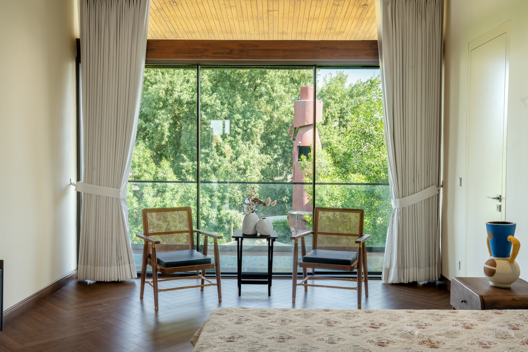 Bedroom of The Holiday Home framing the gulmohar canopy and the restored red kupla chimney beyond full-height glass